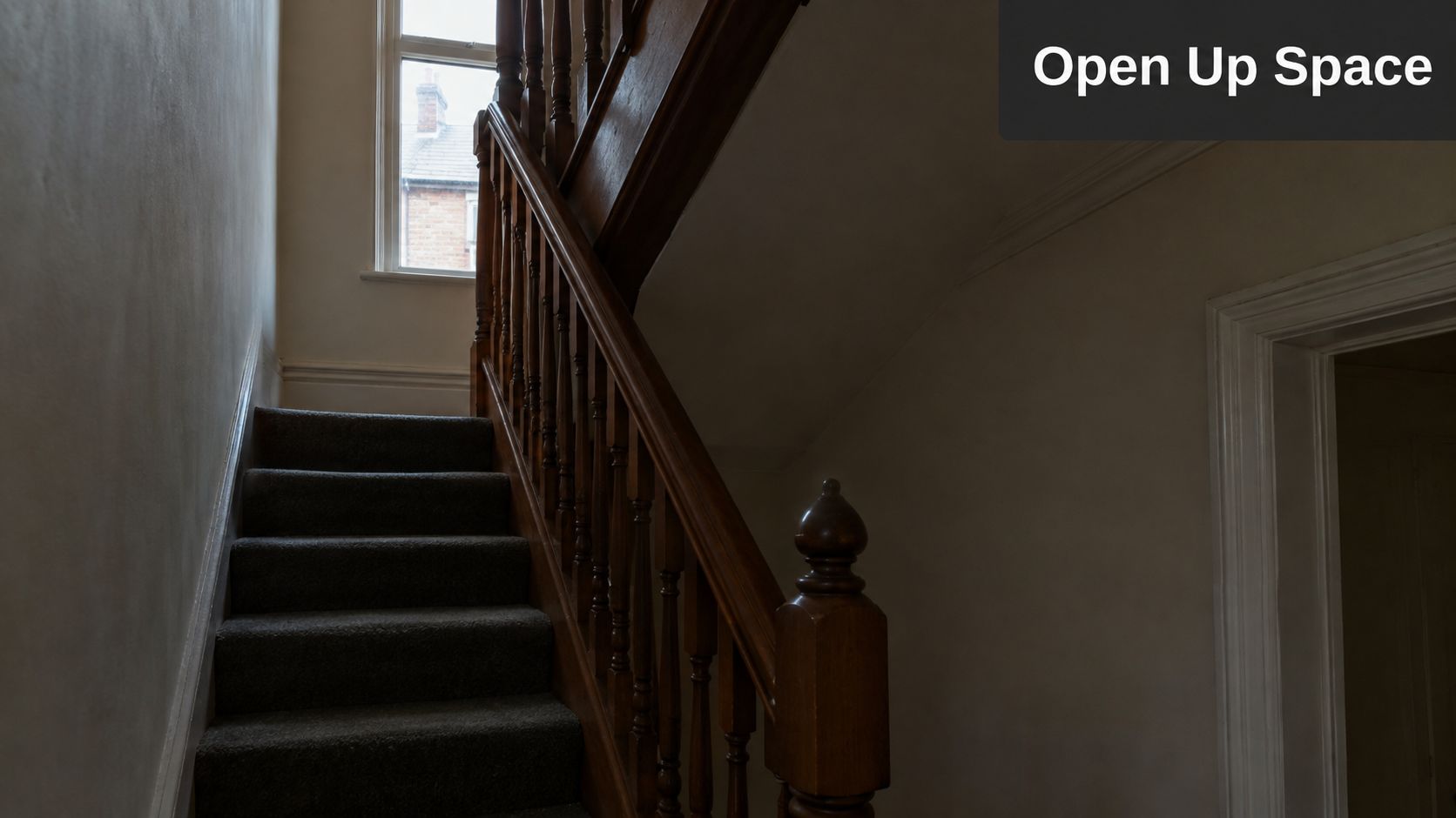 An interior staircase with wooden railings in a dimly lit hallway next to a sunny window